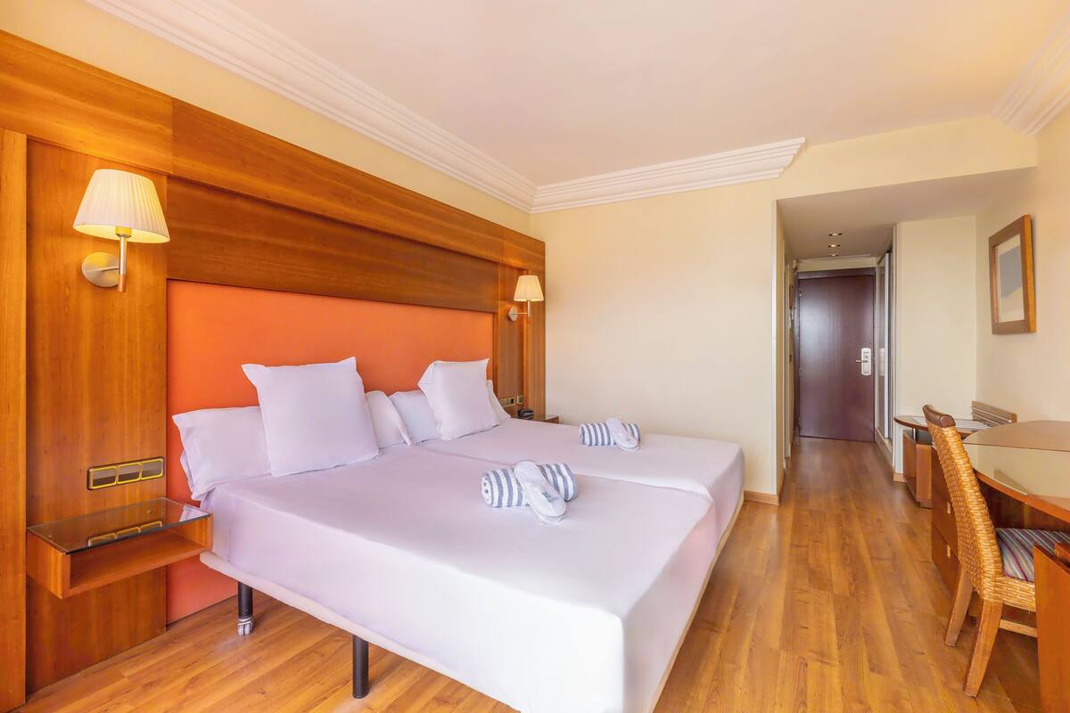 Habitación de hotel con dos camas, cabeceros de madera, una mesita de noche de madera, una mesita auxiliar de vidrio y una mesa de comedor con sillas, además de una puerta que da a otra habitación.