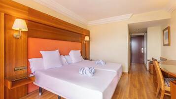 Habitación de hotel con dos camas, cabeceros de madera, una mesita de noche de madera, una mesita auxiliar de vidrio y una mesa de comedor con sillas, además de una puerta que da a otra habitación.