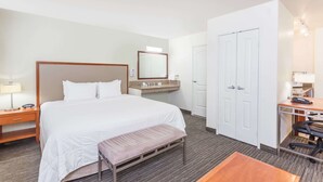 A hotel room with a large bed, a desk, a chair, a bench, a mirror, a vanity with a sink, and a wardrobe. - Hawthorn Suites by Wyndham Victorville (Victorville)