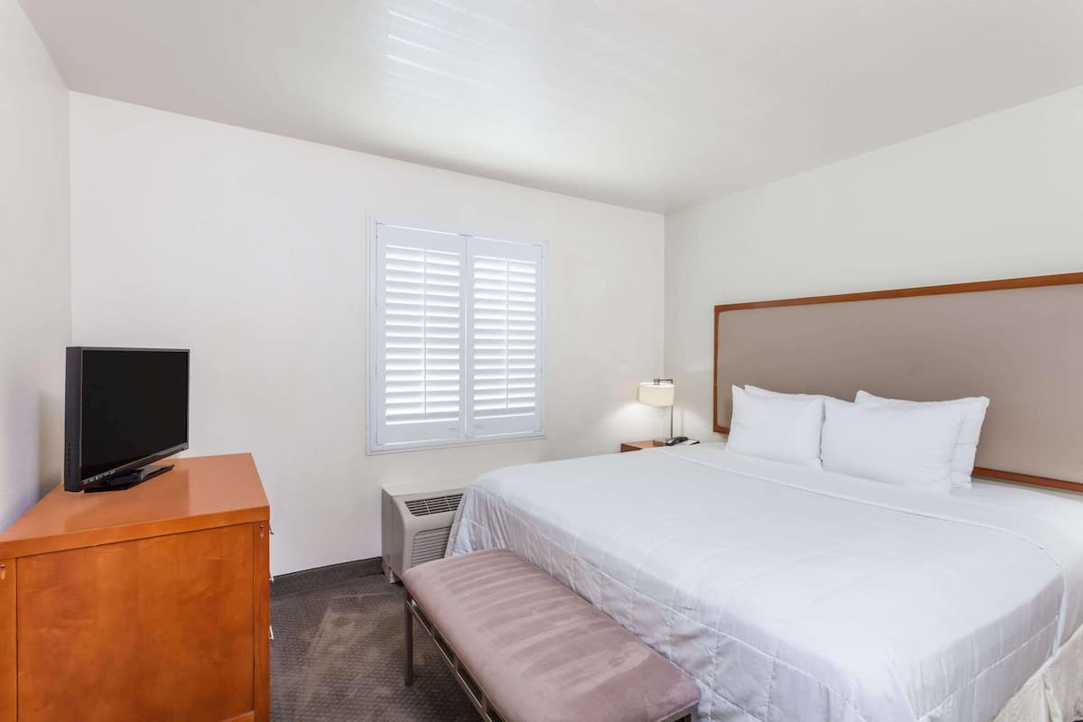 A hotel room with a bed, a TV on a wooden cabinet, a bench, a window with blinds, and a bedside lamp.