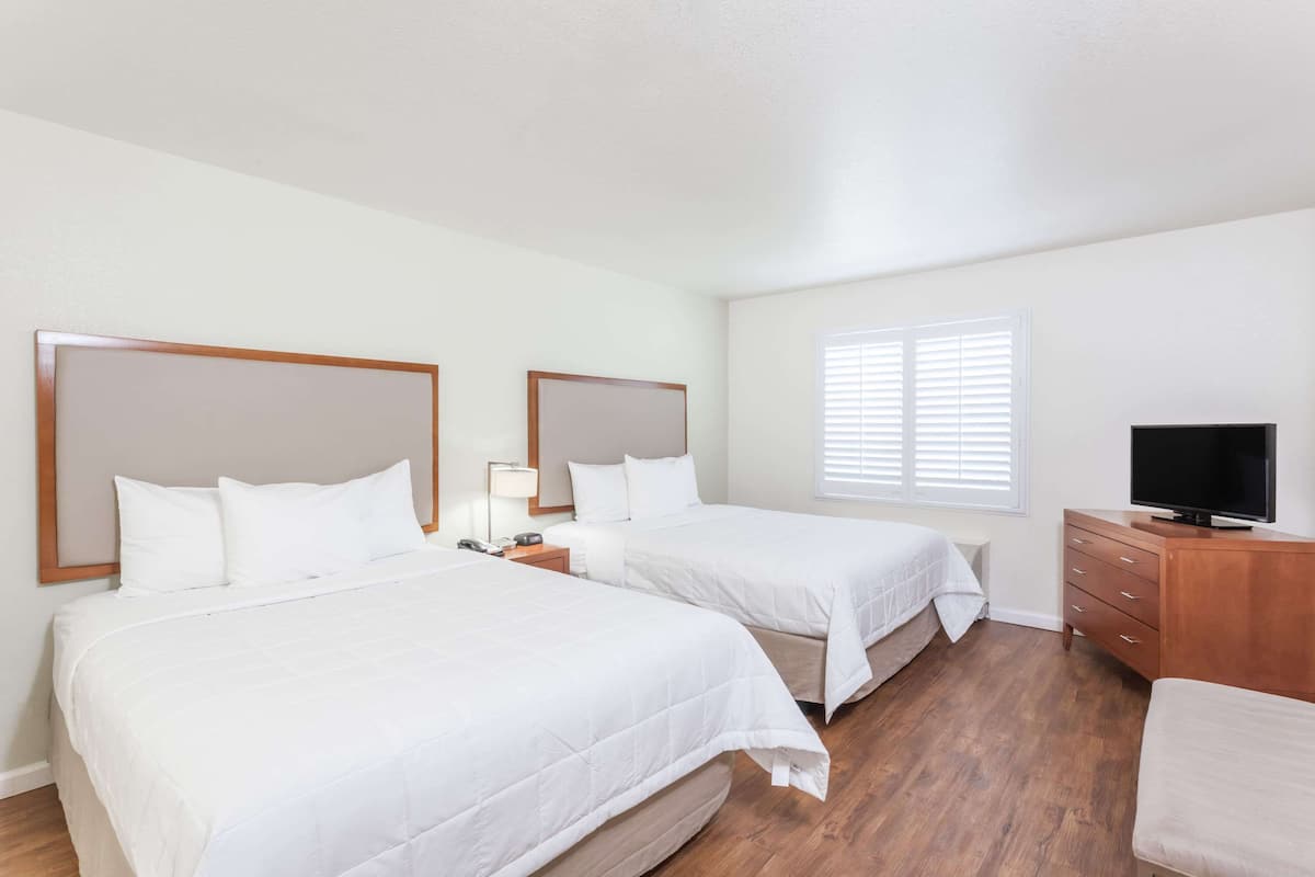 A hotel room with two beds, a TV, a wooden dresser, and a window with blinds.