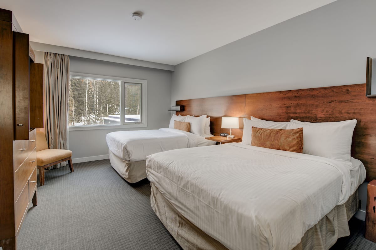 Une chambre d’hôtel avec deux lits, une tête de lit en bois, une fenêtre donnant sur des arbres et un bureau avec une lampe.