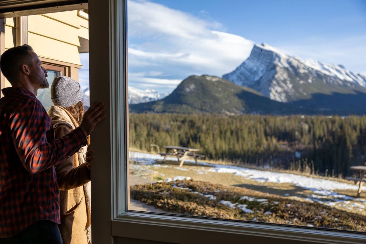 Une personne tenant un enfant près d’une fenêtre, avec vue sur une montagne et une forêt.