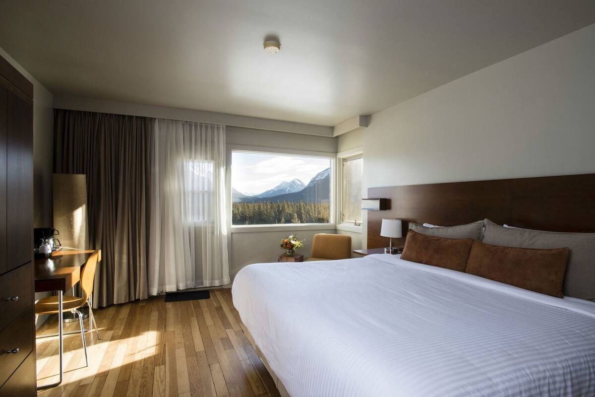 Une chambre d’hôtel avec un lit, un bureau et une chaise, une lampe, et une vue sur les montagnes par la fenêtre.