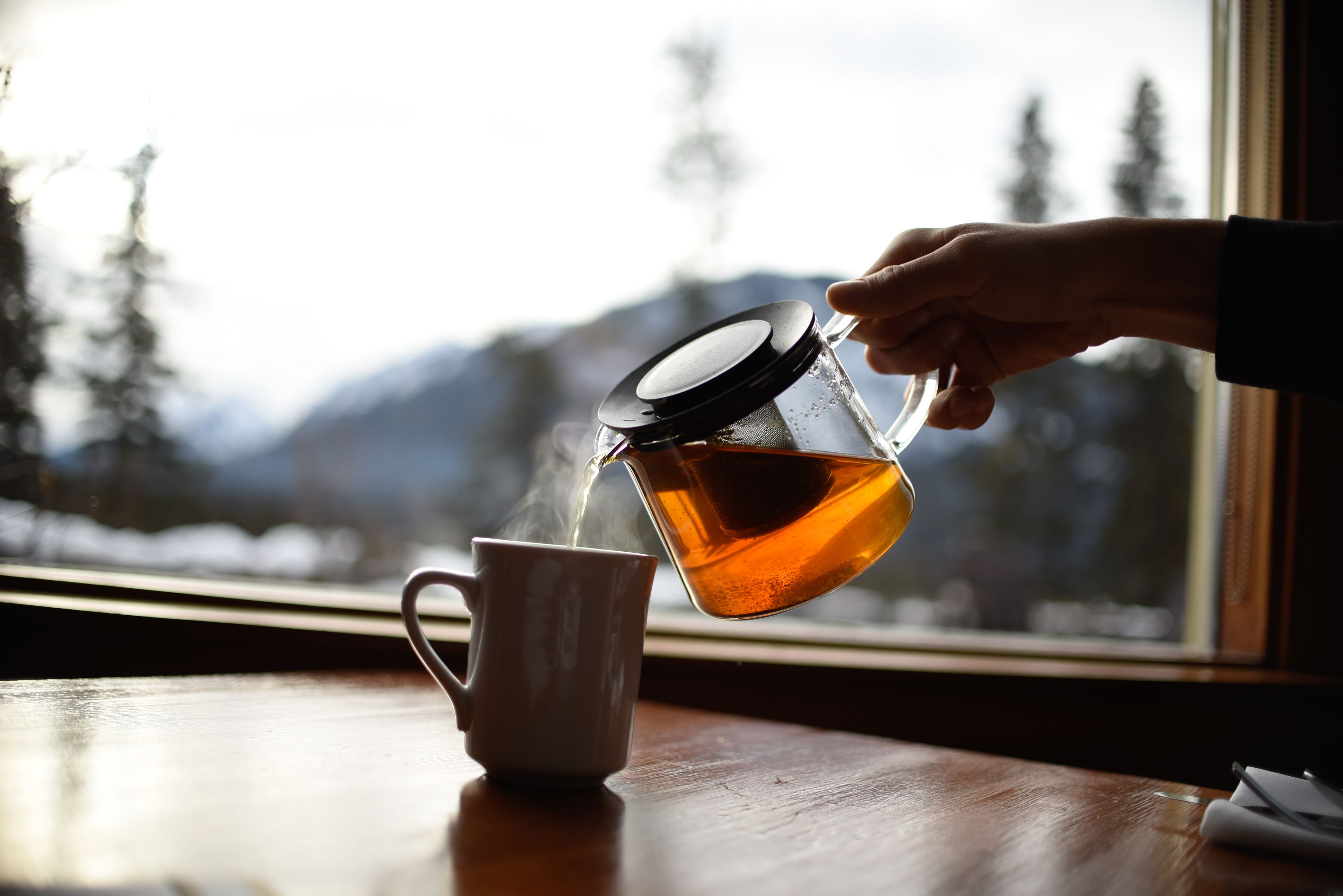 Una mano sirve té en una taza blanca sobre una mesa de madera junto a una ventana con vistas a una montaña nevada.