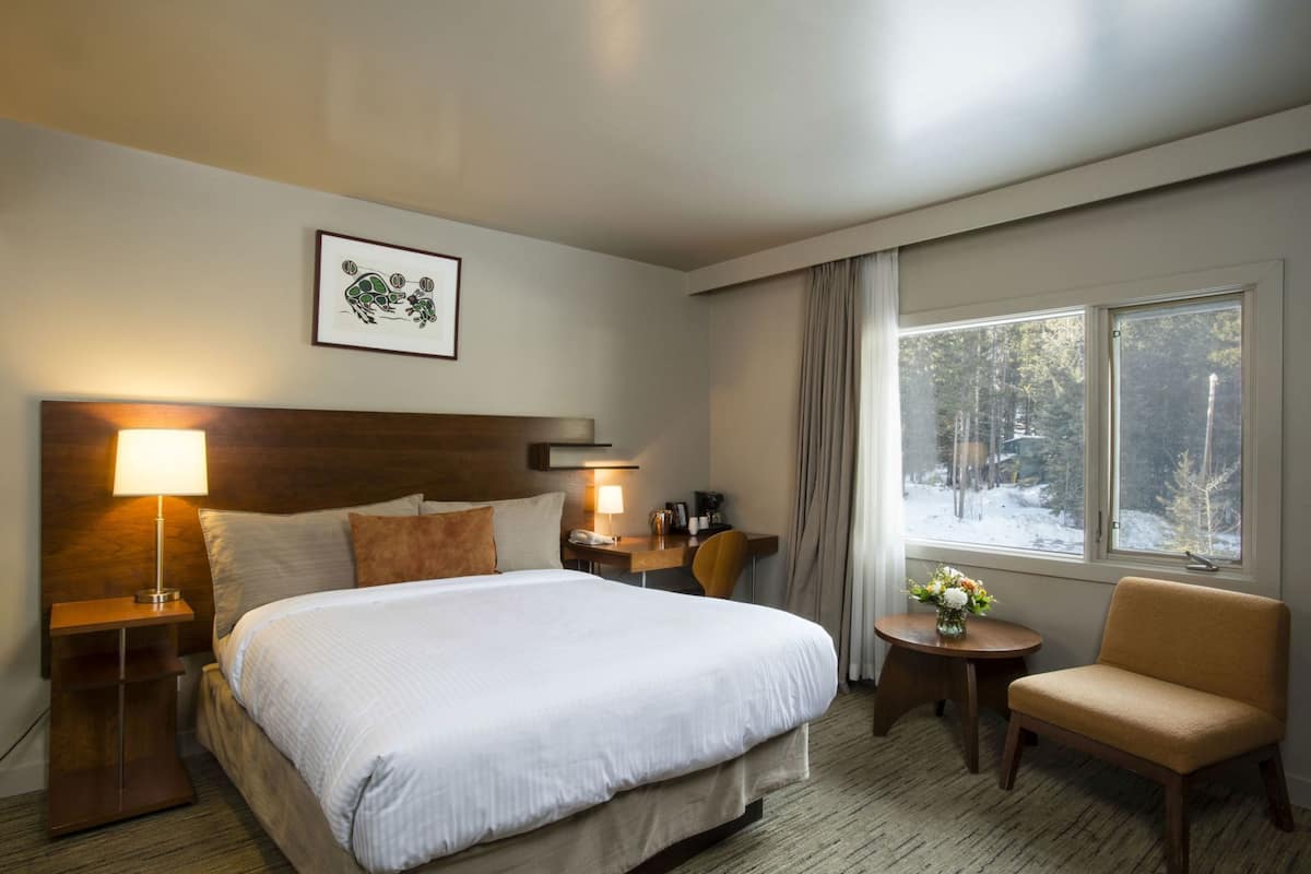 Une chambre d’hôtel avec un grand lit, des lampes de chevet, une petite table avec un vase, une chaise et une fenêtre donnant sur des arbres.
