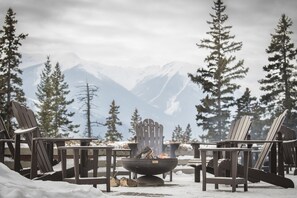 A snow-covered outdoor seating area with Adirondack chairs and a fire pit, surrounded by snow and pine trees, with mountains in the background.