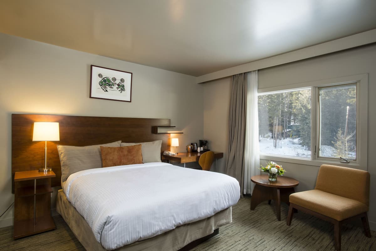 Une chambre d’hôtel avec un grand lit, un bureau, une chaise, une table de chevet, une lampe, un tableau encadré au mur et une fenêtre donnant sur des arbres.