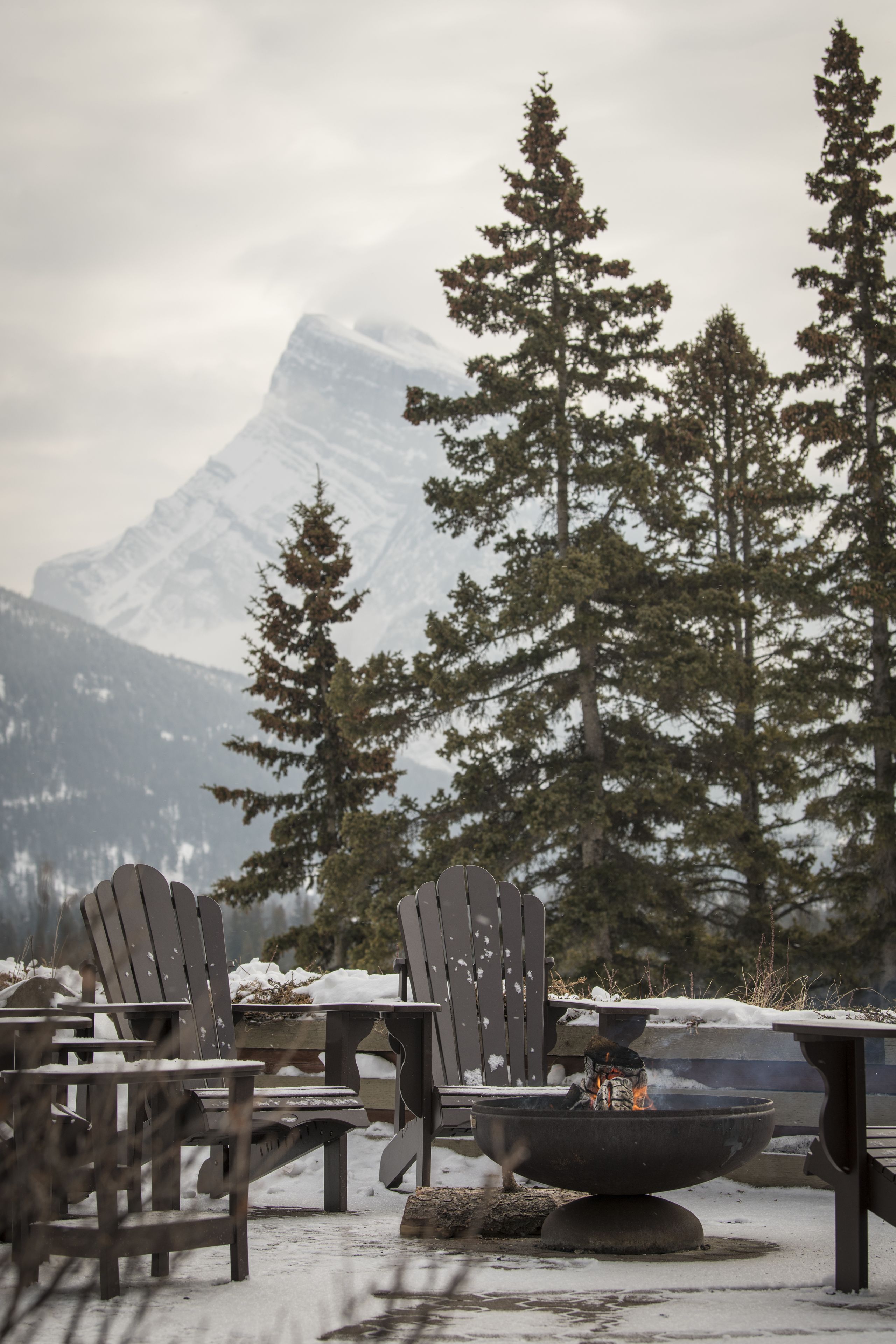 Un espace extérieur aménagé avec des sièges, recouvert de neige, comprenant des fauteuils Adirondack et un foyer, le tout mis en valeur par un paysage de montagnes enneigées et de grands arbres à feuilles persistantes.