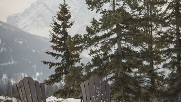 Un espace extérieur aménagé avec des sièges, recouvert de neige, comprenant des fauteuils Adirondack et un foyer, le tout mis en valeur par un paysage de montagnes enneigées et de grands arbres à feuilles persistantes.