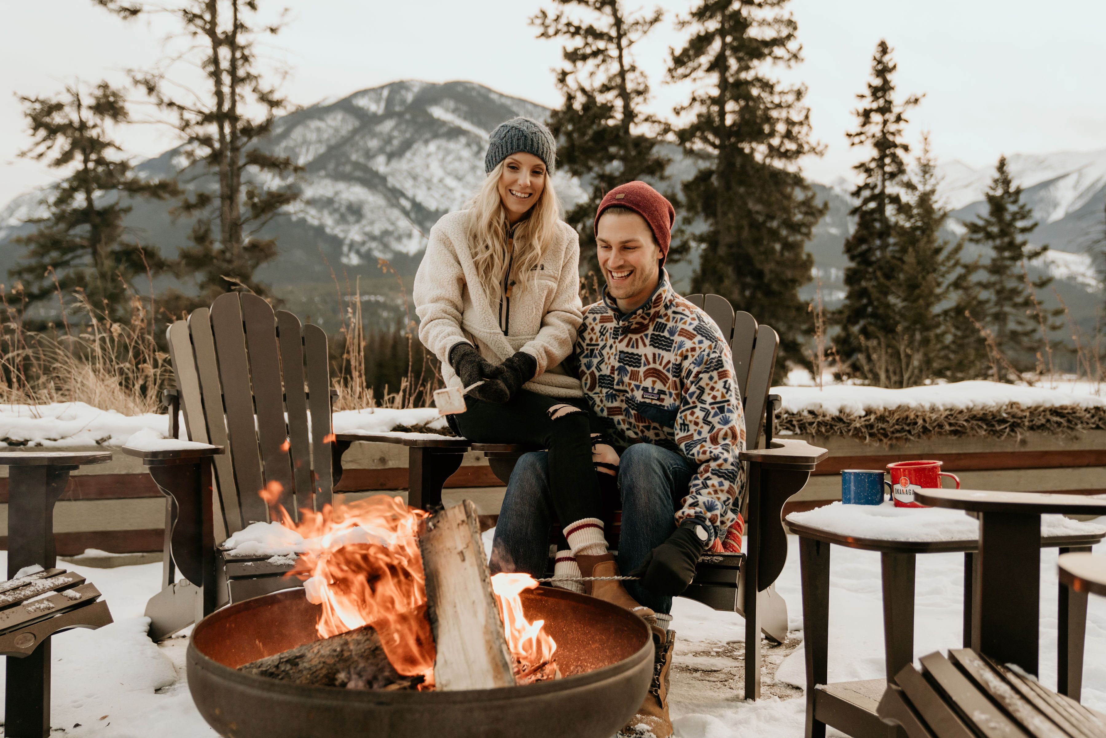 Two people sitting on Adirondack chairs around a campfire in a snowy outdoor setting with mountains in the background.