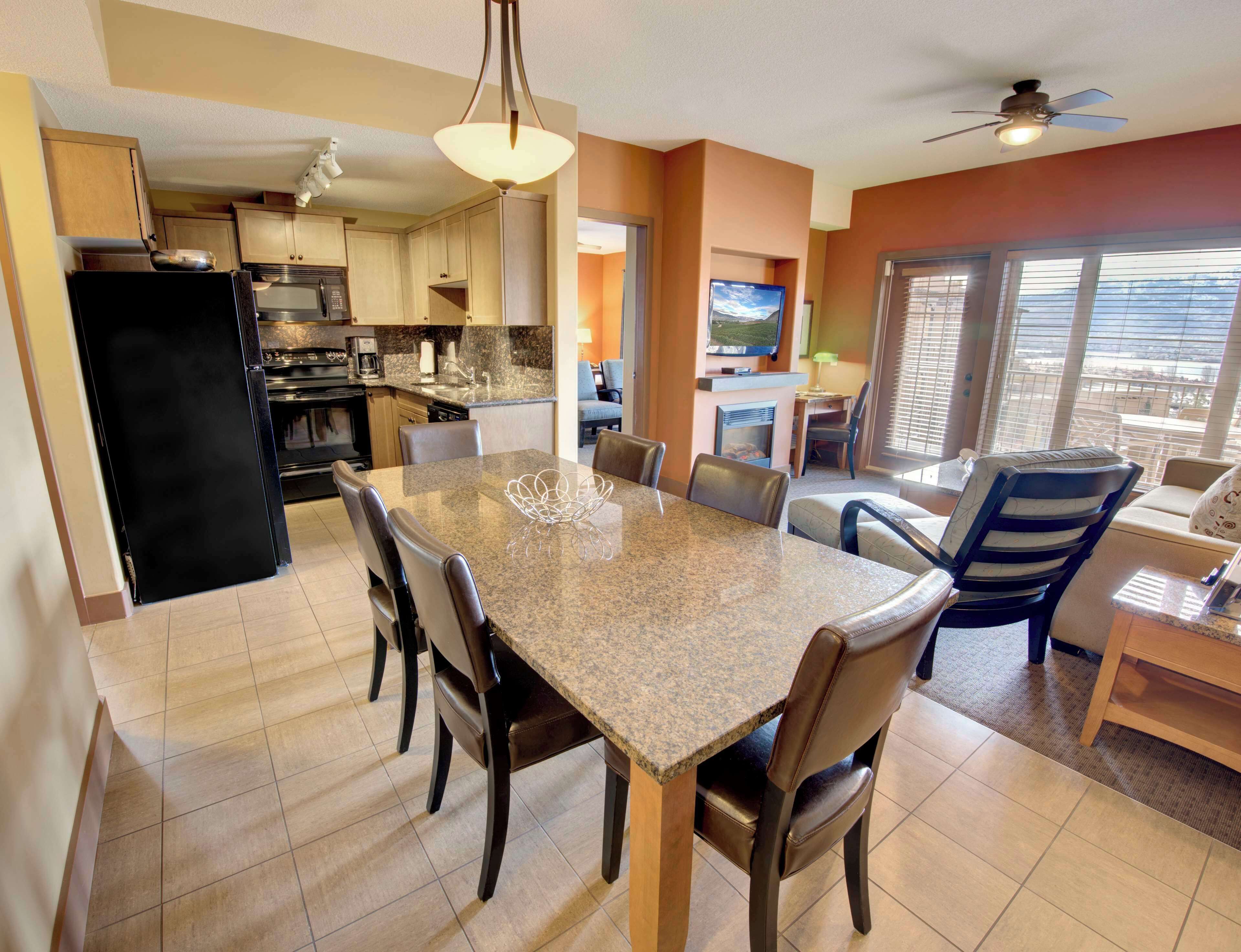 A modern kitchen with granite countertops, a black refrigerator, and a dining area with a granite table and leather chairs.