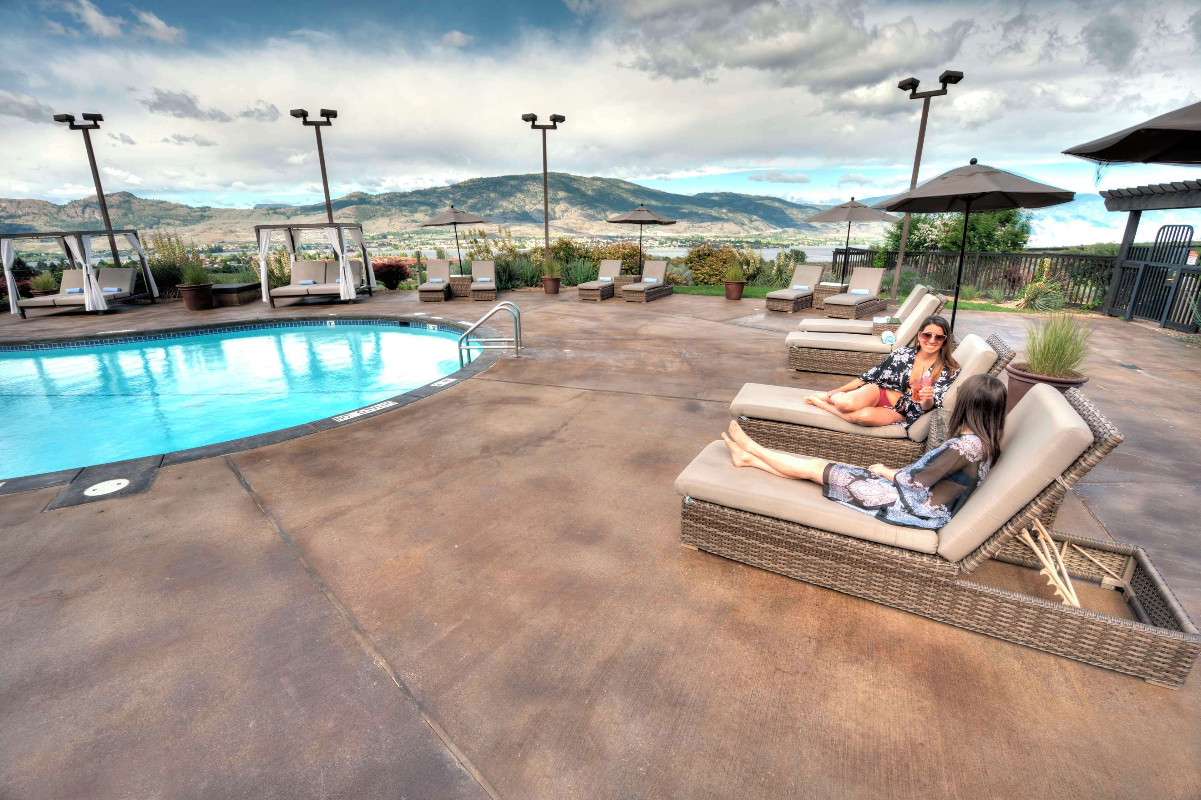 A poolside area with lounge chairs and umbrellas, a swimming pool, and a mountainous backdrop.