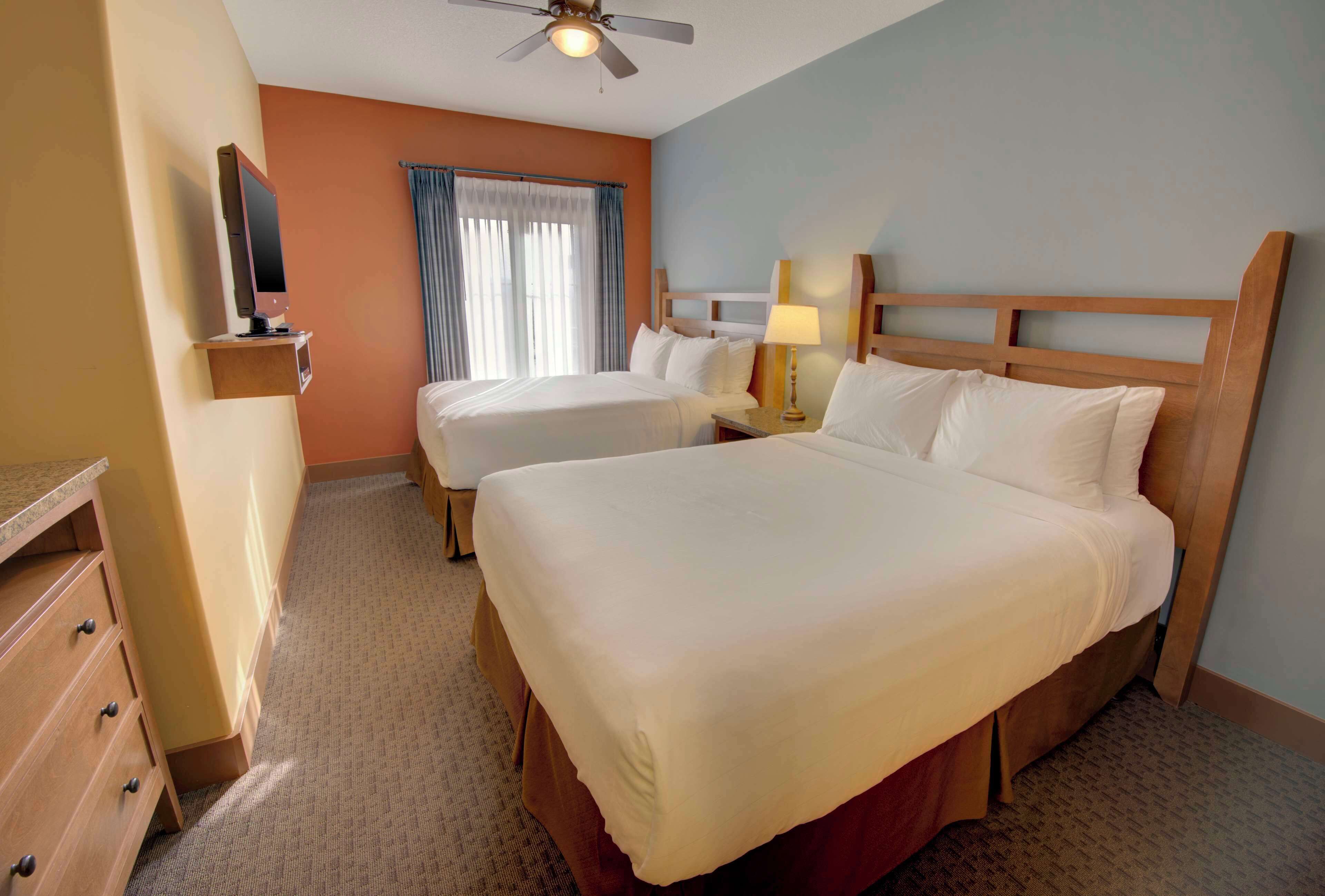A hotel room with two beds, a TV on a stand, a ceiling fan, and a window with curtains.