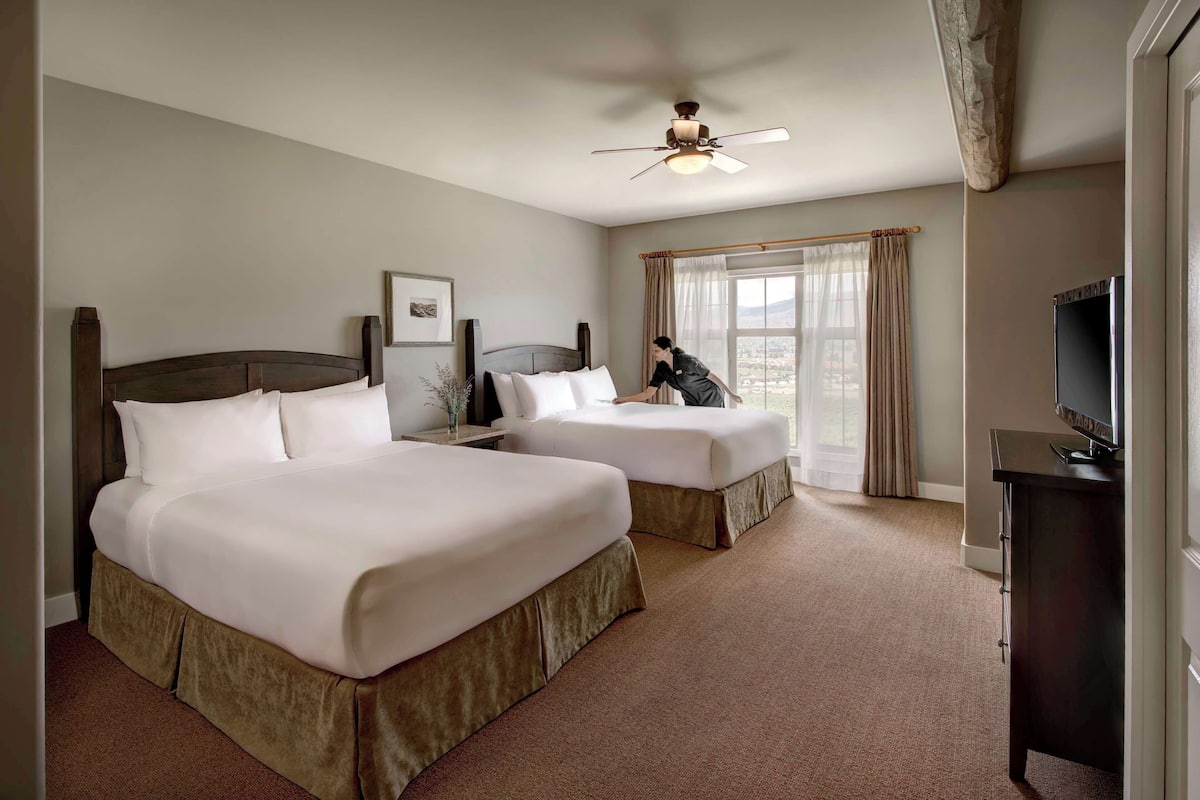 A hotel room with two beds, a ceiling fan, a TV, and a window with curtains.