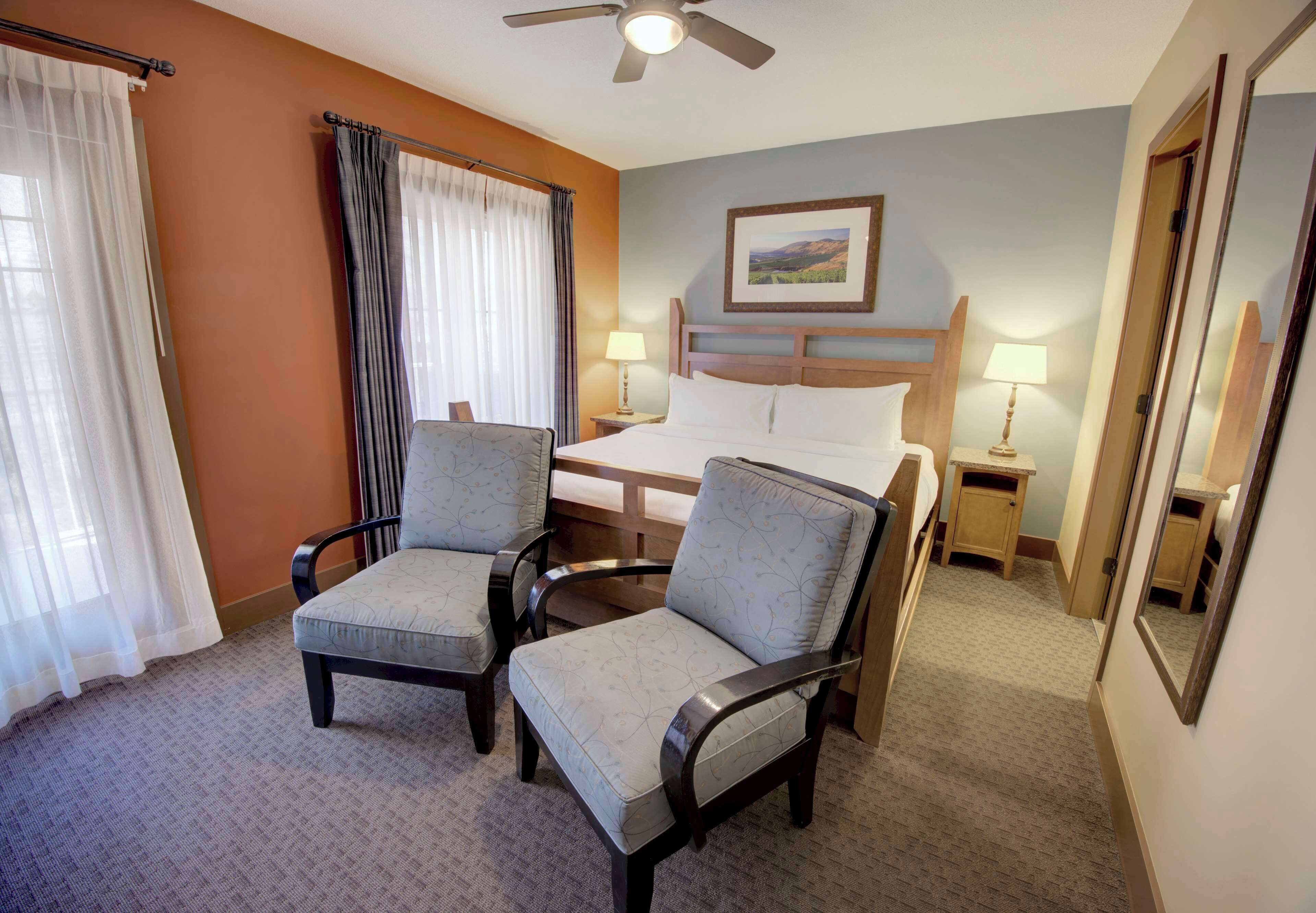 A hotel room with a bed, two armchairs, a nightstand with a lamp, a ceiling fan, and a framed picture on the wall.