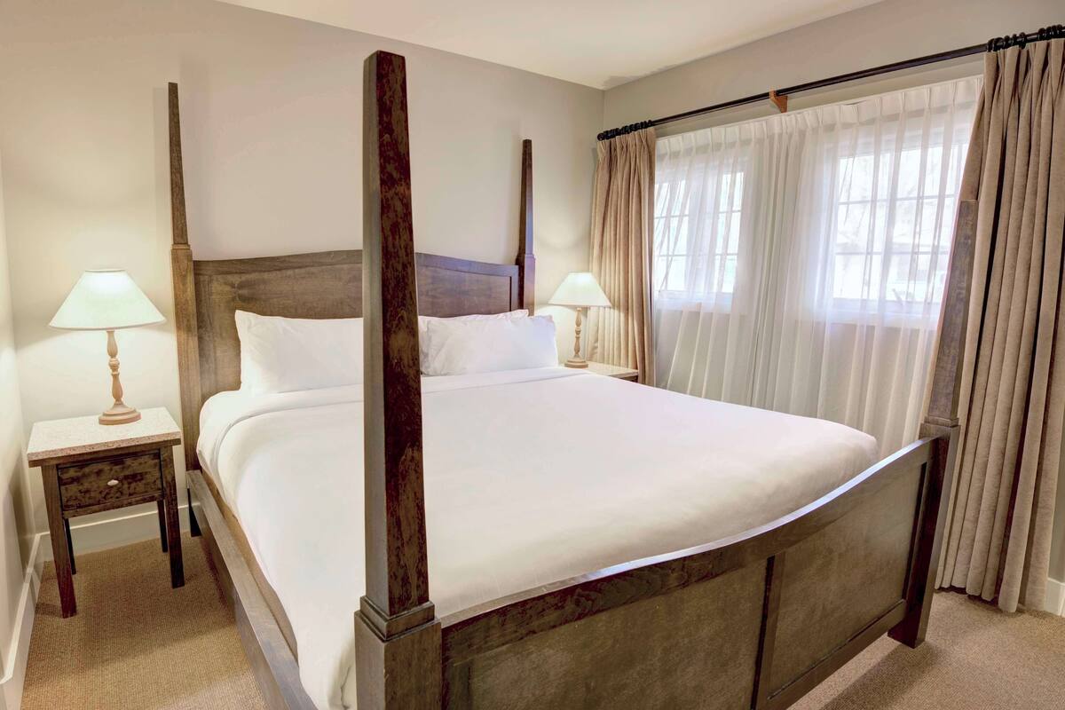 A four-poster bed with white linens, two bedside tables with lamps, and a window with sheer curtains.