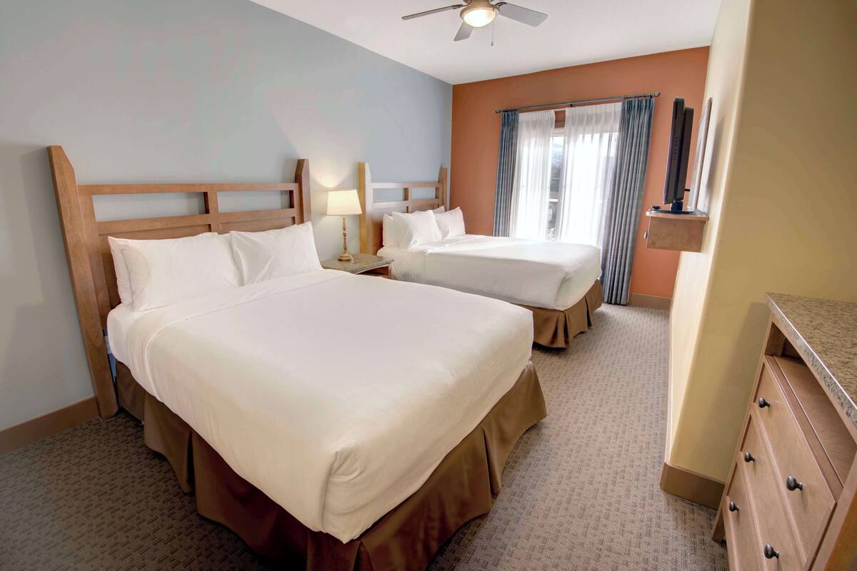 A hotel room with two beds, a ceiling fan, a window with curtains, and a wooden dresser.