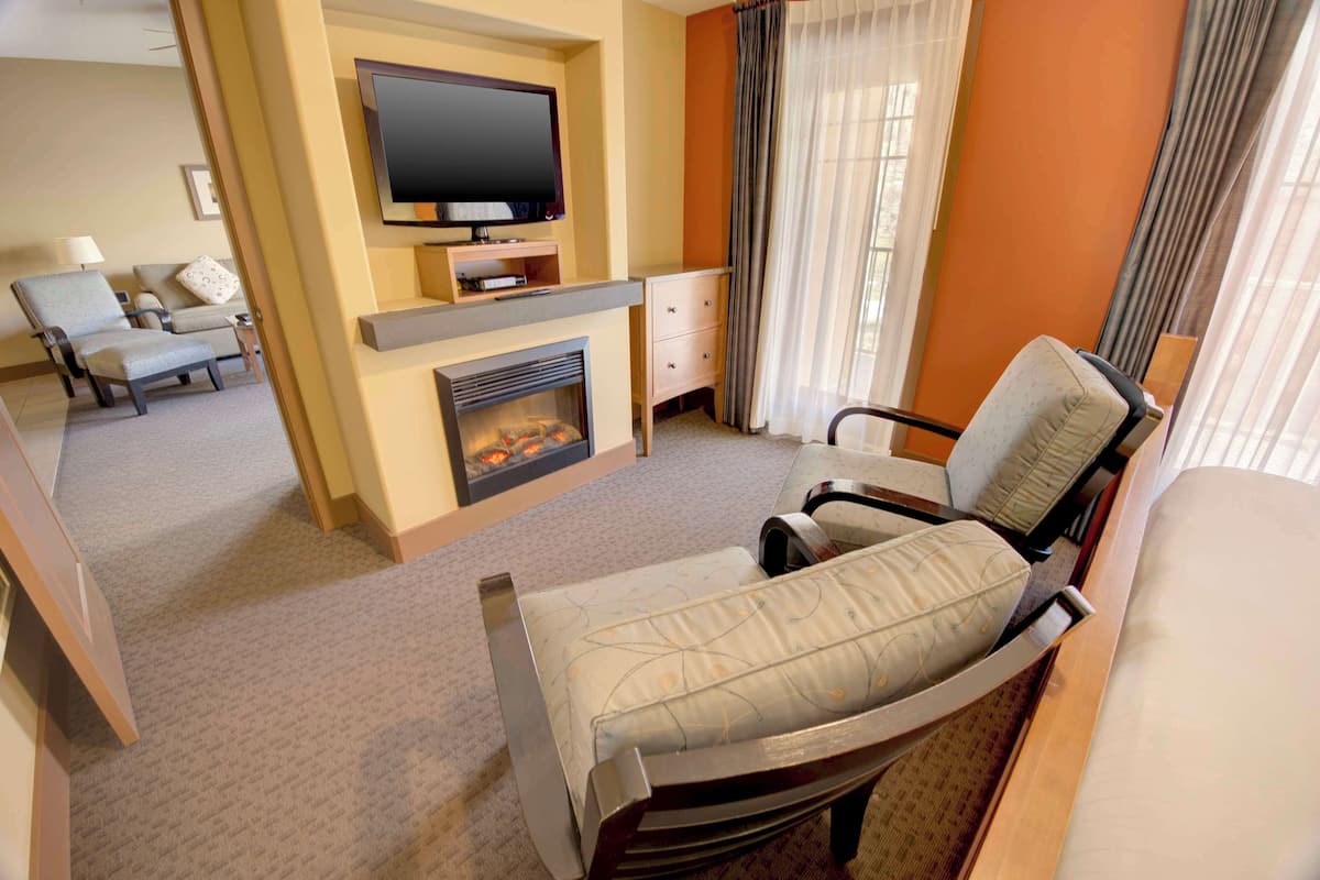 A hotel room with a flat-screen TV mounted above a fireplace, a sofa, a chair, and a bed.