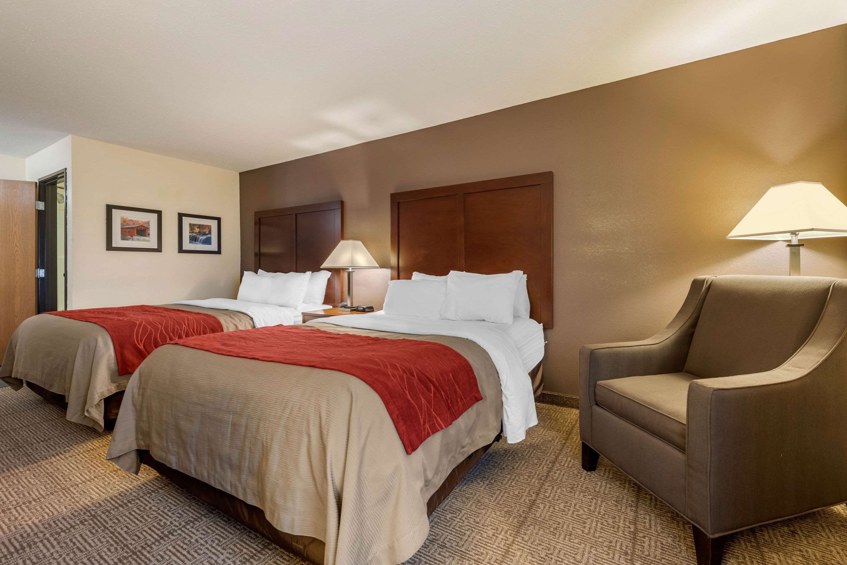 A hotel room with two beds, a sitting area with a chair, a lamp, and two framed pictures on the wall.