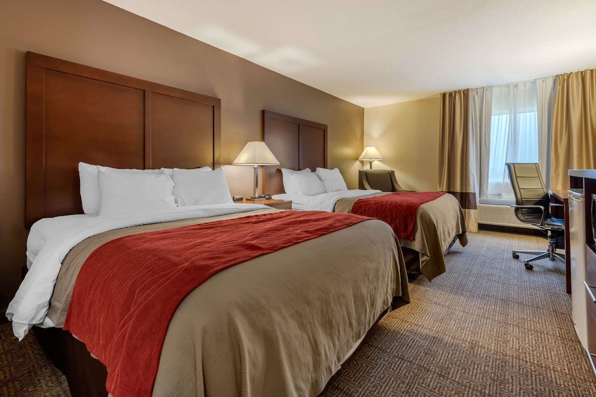 A hotel room with two beds, each with a red and beige bedspread, two bedside lamps, a desk with a chair, and a large window with curtains.