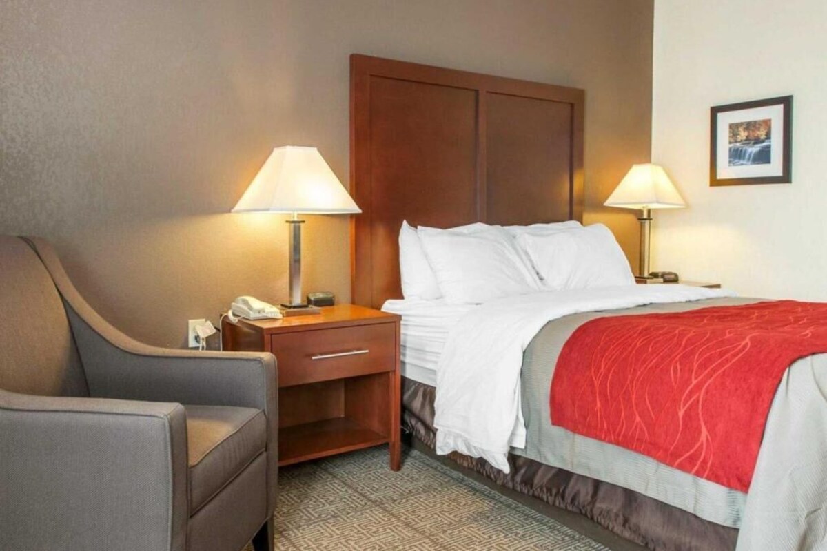 Habitación de hotel con cama, dos lámparas de noche, una silla, mesita de noche y un cuadro en la pared.