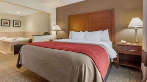 A hotel room with a large bed, a wooden headboard, a bedside table with a lamp, a nightstand, and a bathroom with a bathtub.