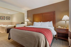A hotel room with a large bed, a wooden headboard, a bedside table with a lamp, a nightstand, and a bathroom with a bathtub.