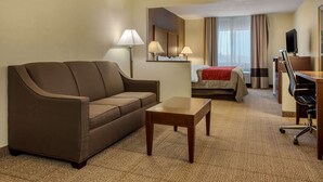 A hotel room with a brown sofa, a wooden coffee table, a bed with a red blanket, a desk with a chair, and a TV on a stand.