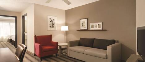 A hotel room with a dining table, a red armchair, a sofa, a lamp, and framed pictures on the wall.