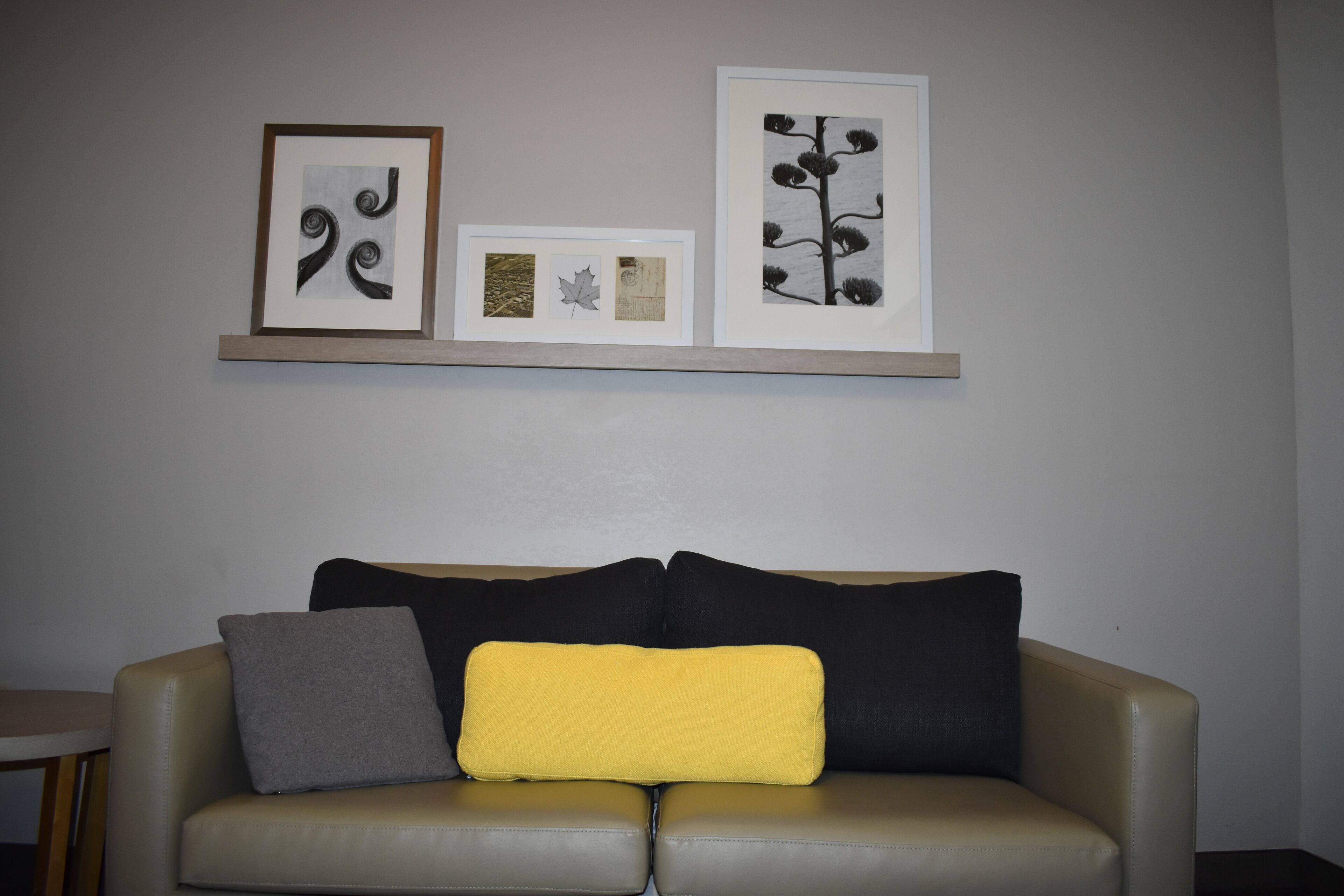 A neutral-toned living room with a beige sofa, two cushions, and a small wooden table. Above the sofa, there are three framed artworks on a wall-mounted shelf.