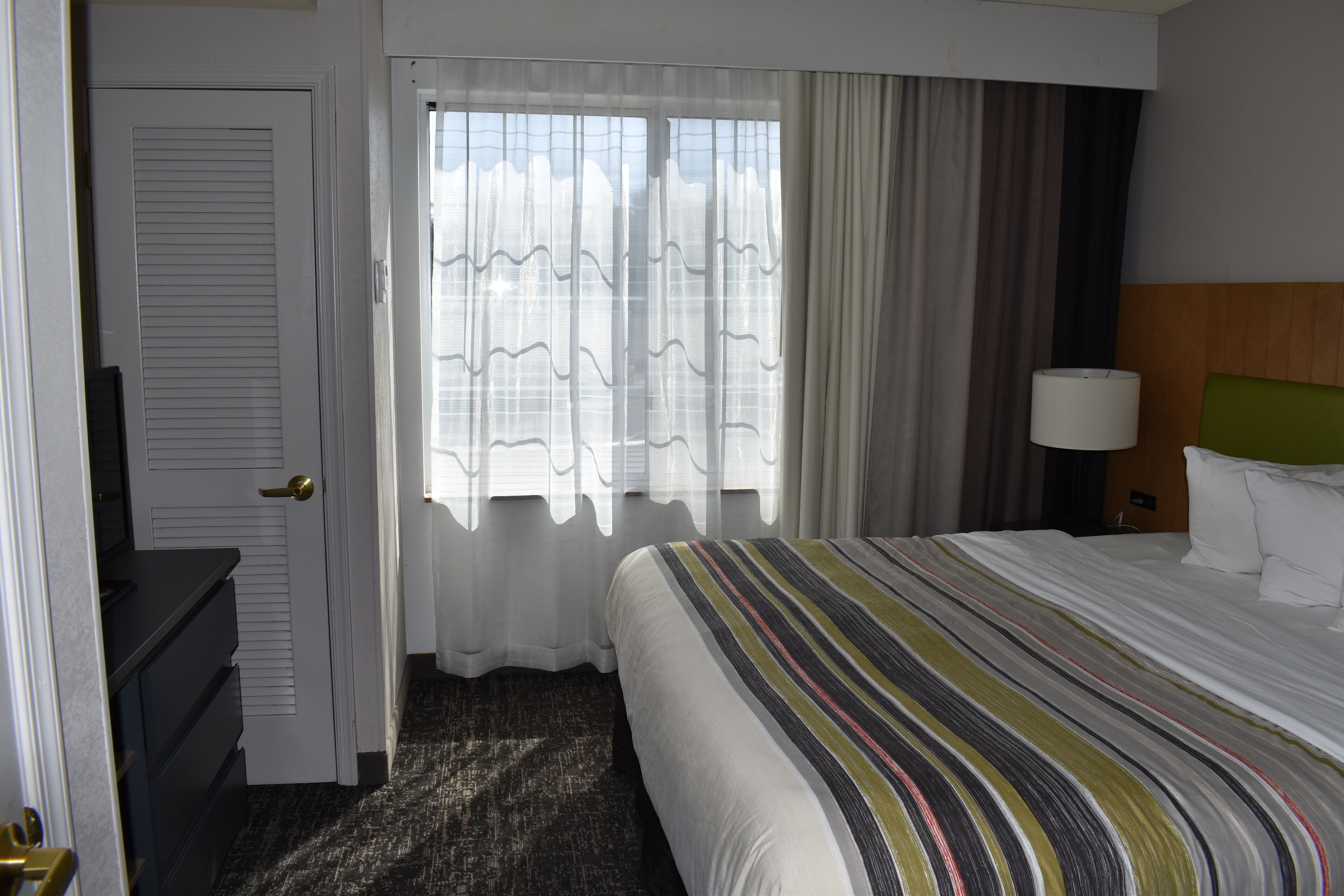 A hotel room with a bed, a nightstand with a lamp, a dresser, and a window with curtains.