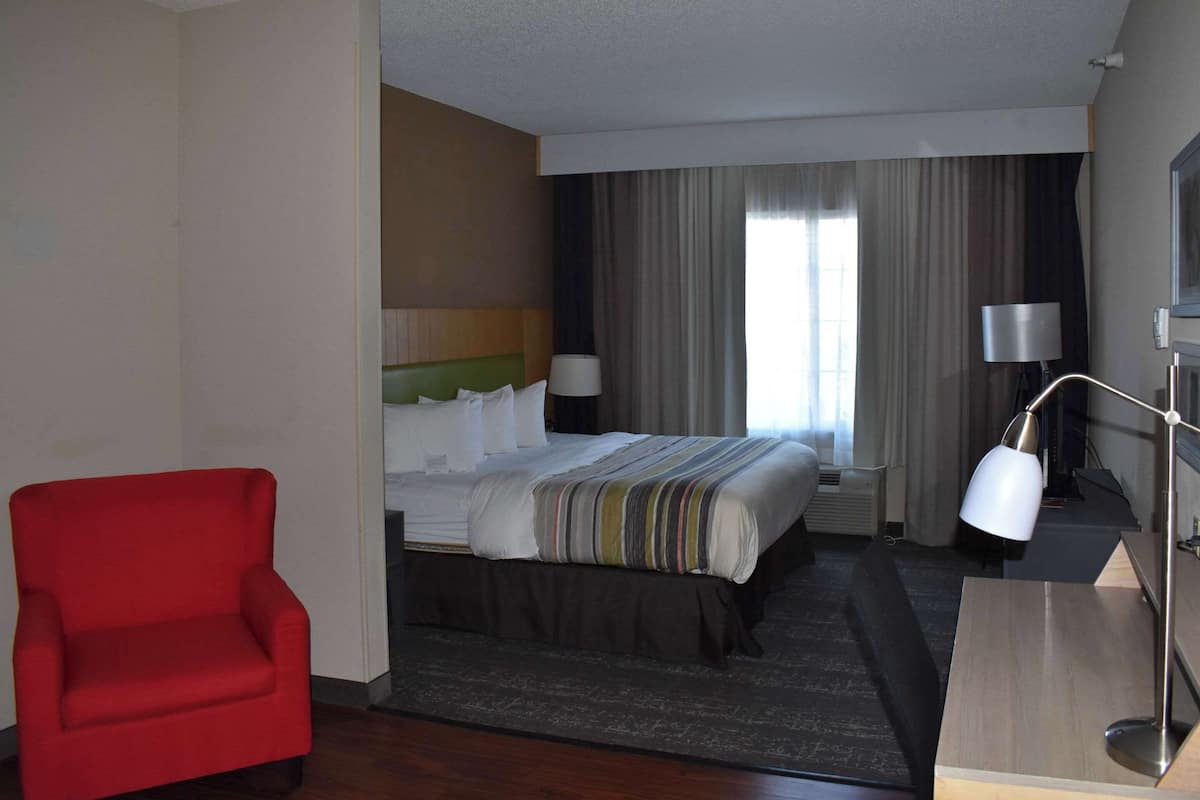 A hotel room with a bed, a red armchair, a desk with a lamp, and a large window with curtains.