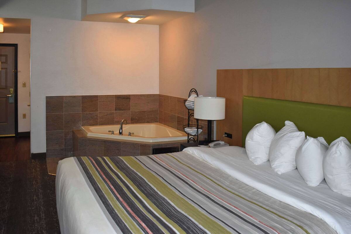 A hotel room with a bed, a large window, a bathroom with a bathtub, and a green headboard.