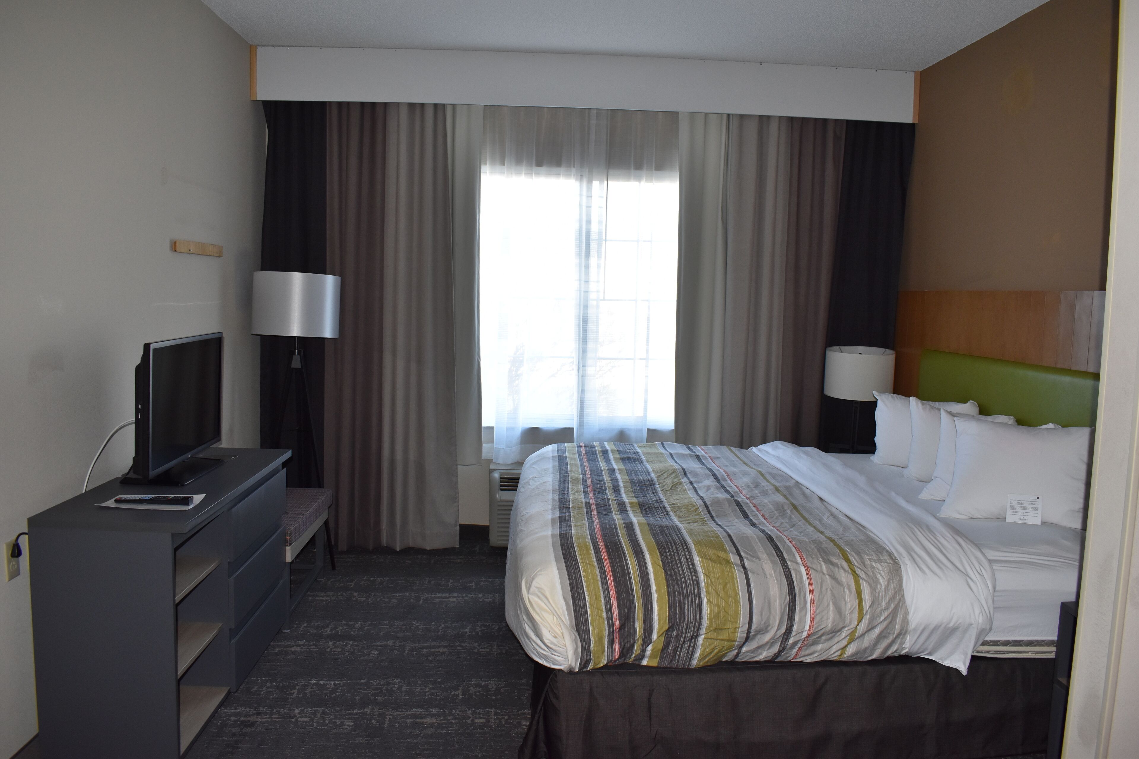 Quarto de hotel com cama, televisão, escrivaninha e janela com cortinas.
