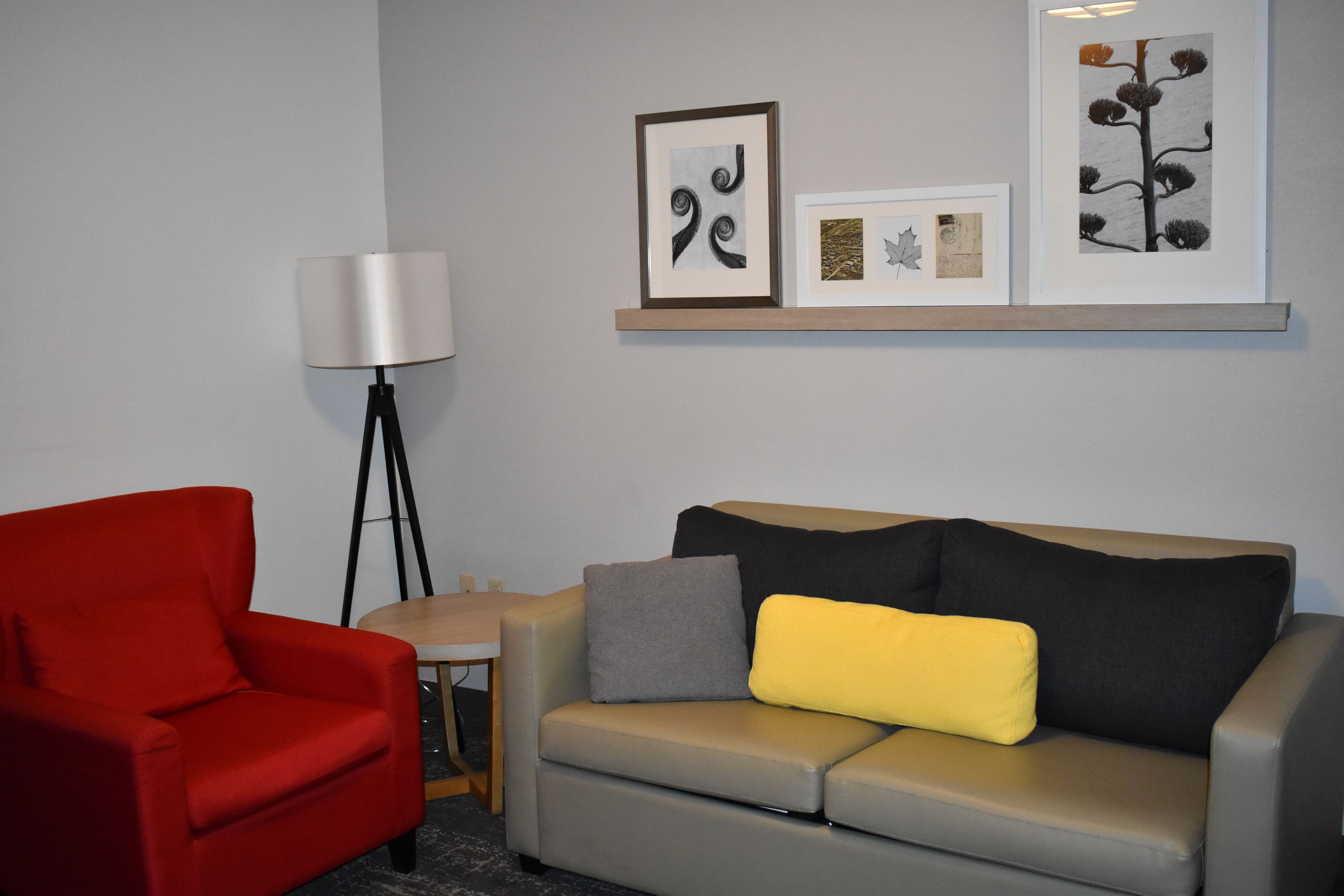 A modern room with a grey sofa, a red armchair, a floor lamp, and a wall-mounted shelf with framed artwork.