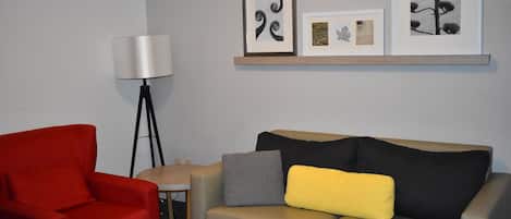 Quarto moderno com sofá cinza, poltrona vermelha, luminária de chão e prateleira com obras de arte emolduradas.