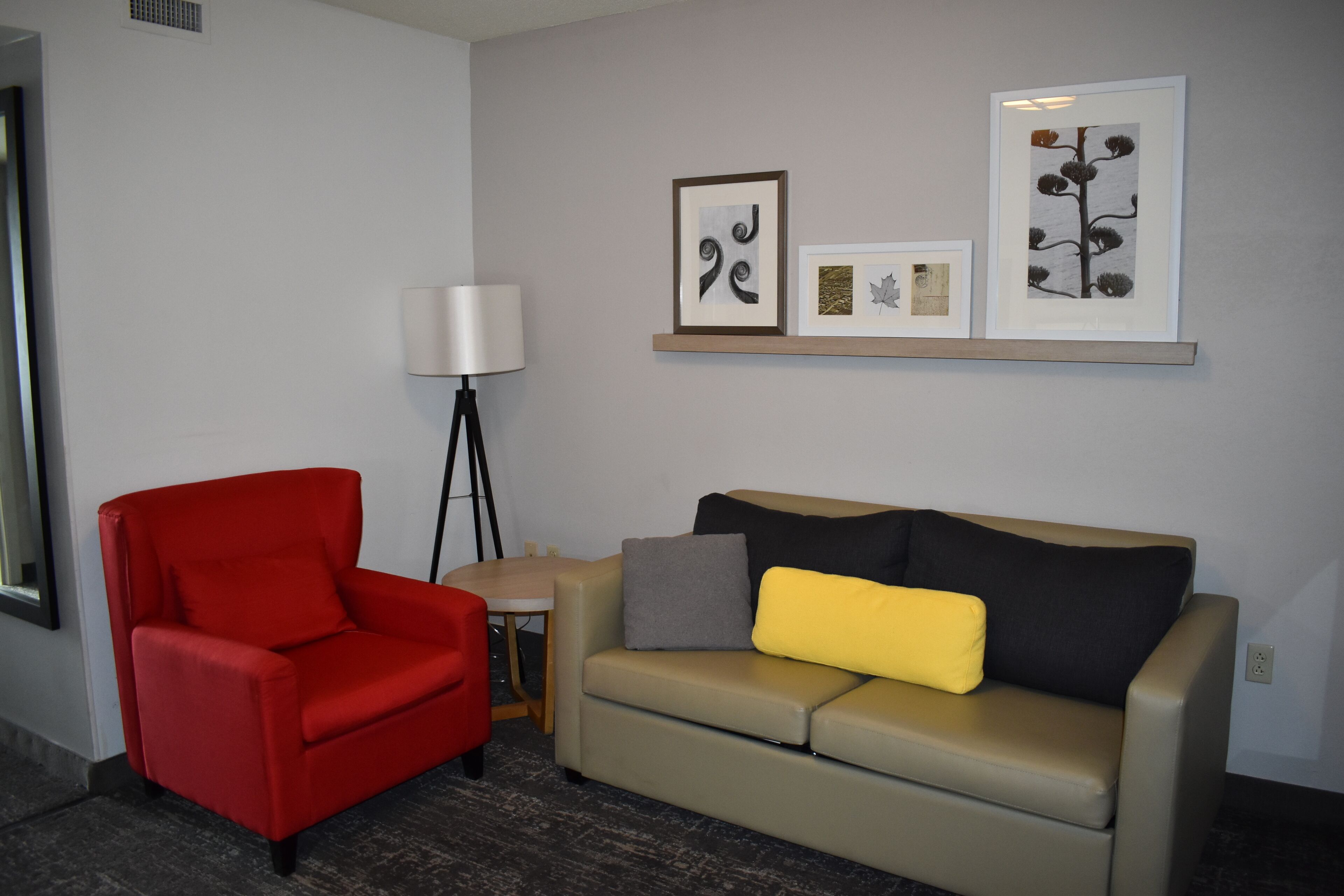 A room with a red chair, a beige sofa with a yellow pillow, a floor lamp, and framed artwork on the wall.