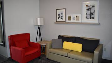 A room with a red chair, a beige sofa with a yellow pillow, a floor lamp, and framed artwork on the wall.