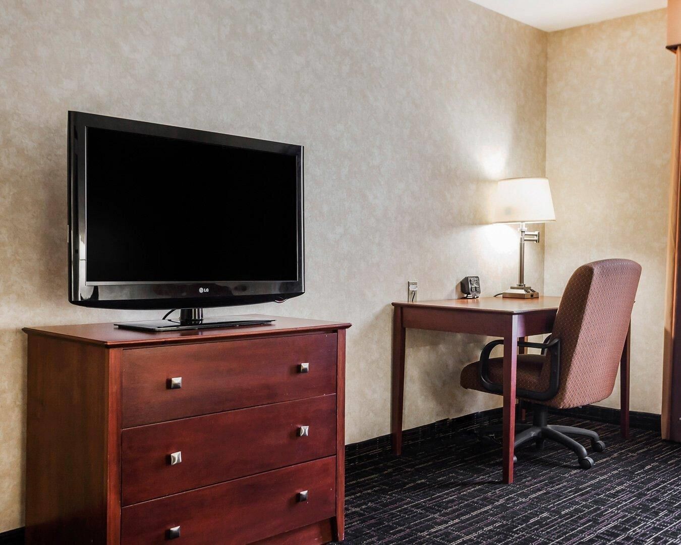 Habitación de hotel con televisión de pantalla plana sobre un aparador de madera, un escritorio con lámpara y una silla.