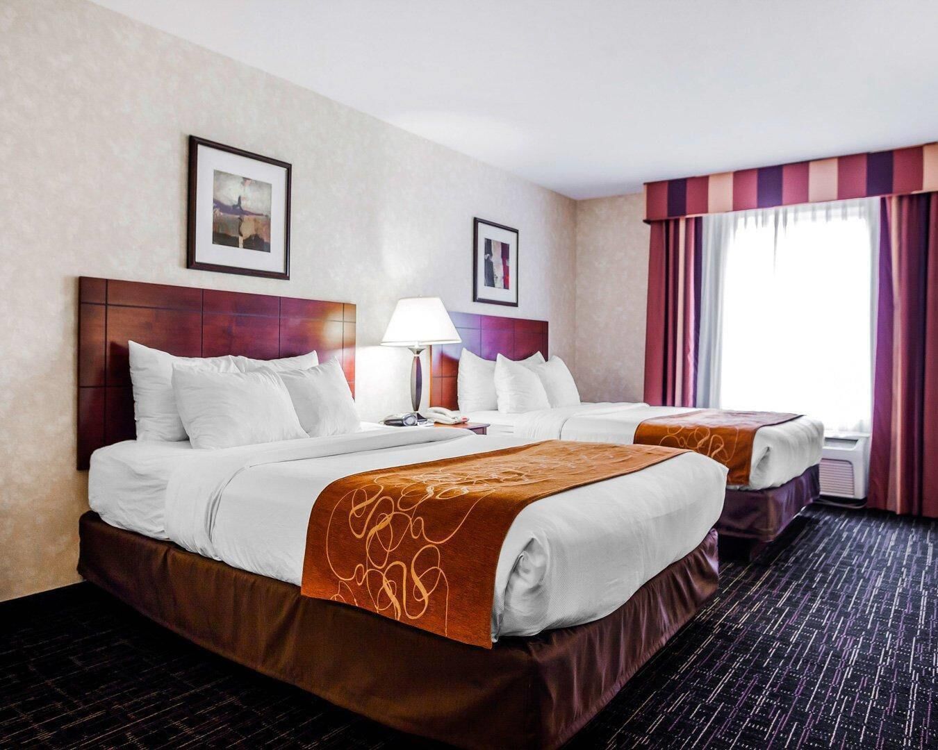 A hotel room with two beds, a nightstand with a lamp, a window with curtains, and framed pictures on the wall.