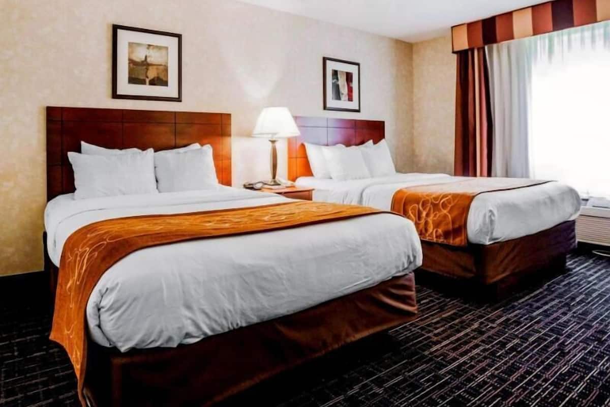 A hotel room with two beds, a nightstand, a lamp, and a window with curtains.