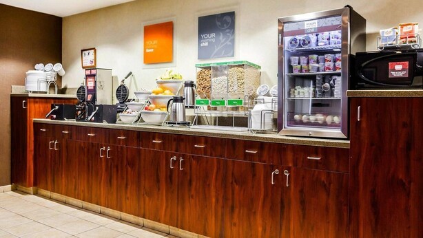 Espaço de café da manhã de hotel, com balcão de madeira, máquina de café, aparelho de waffle e geladeira abastecida com bebidas e lanches.