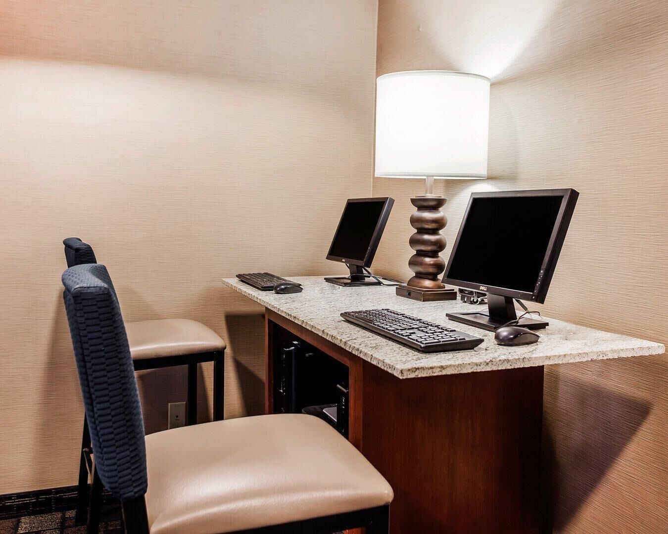 Una habitación de hotel con un escritorio, dos monitores, un teclado, un ratón, una lámpara y una silla.