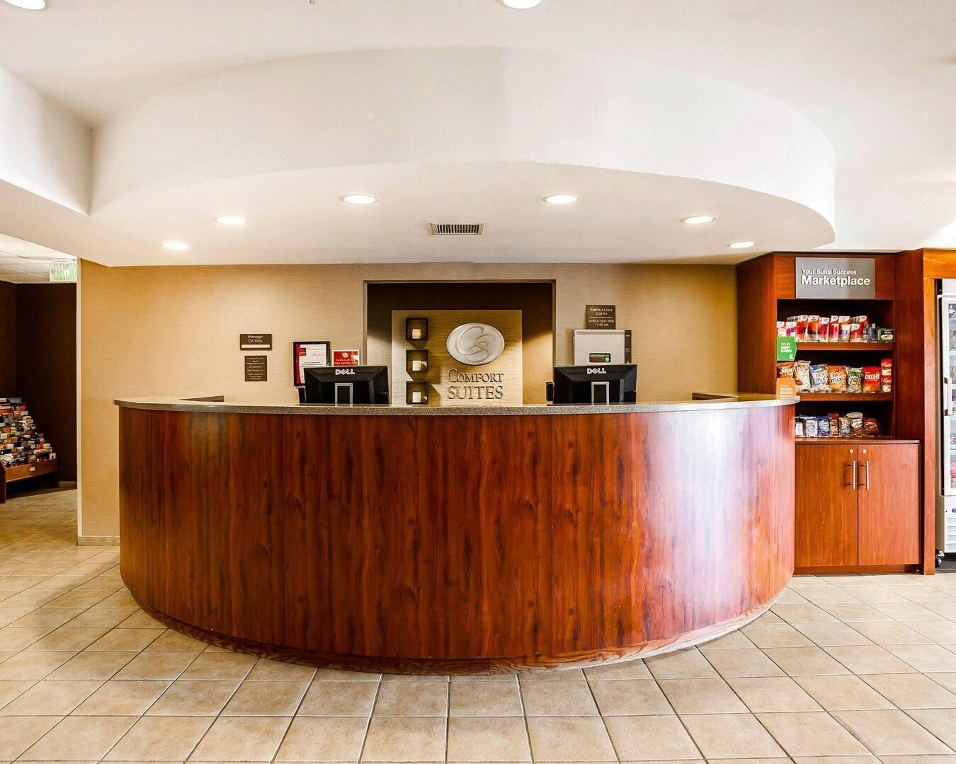 A hotel reception area with a wooden reception desk, a sign for 'Comfort Suites', and a kitchenette with a refrigerator and shelves of snacks.