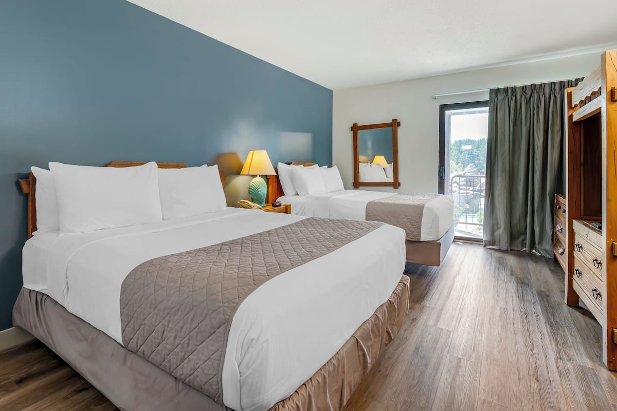 Habitación de hotel con dos camas, un aparador de madera, una mesita de noche con lámpara y un ventanal con cortinas.