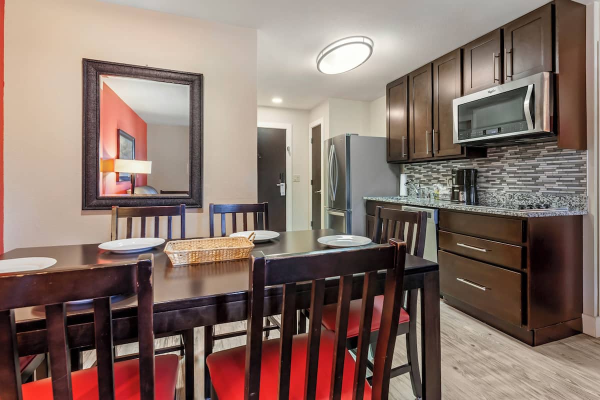 Un comedor con una mesa de madera oscura, sillas rojas y un espejo grande. Hay una cocina con armarios de madera oscura, un microondas de acero inoxidable y un salpicadero de azulejos.