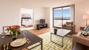 A hotel room with a dining table set for four, a sofa, a TV stand, and a large window offering a city view. - Oakwood Crystal City (Arlington)