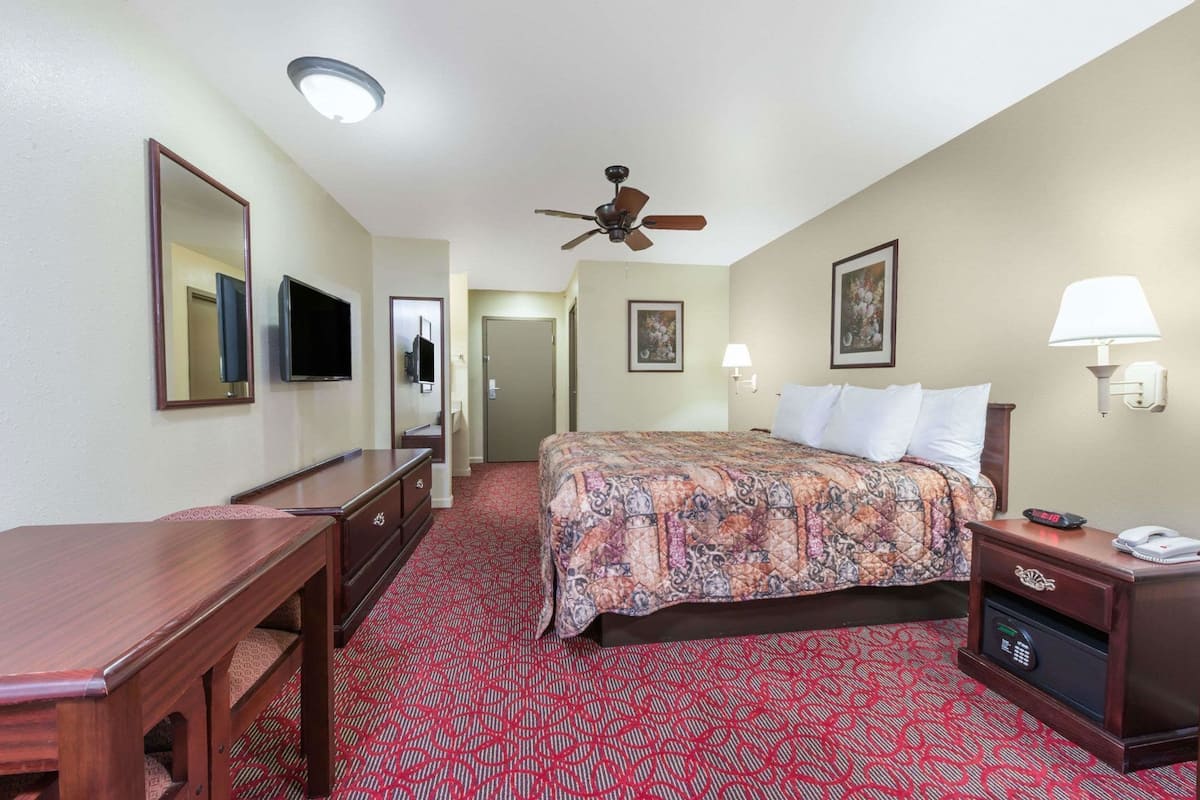 A hotel room with a bed, a desk, a television, a ceiling fan, and a red carpeted floor.