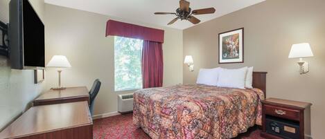 Una habitación de hotel con cama, escritorio, silla, televisor, ventilador de techo, ventana con cortinas y mesita de noche con lámpara.